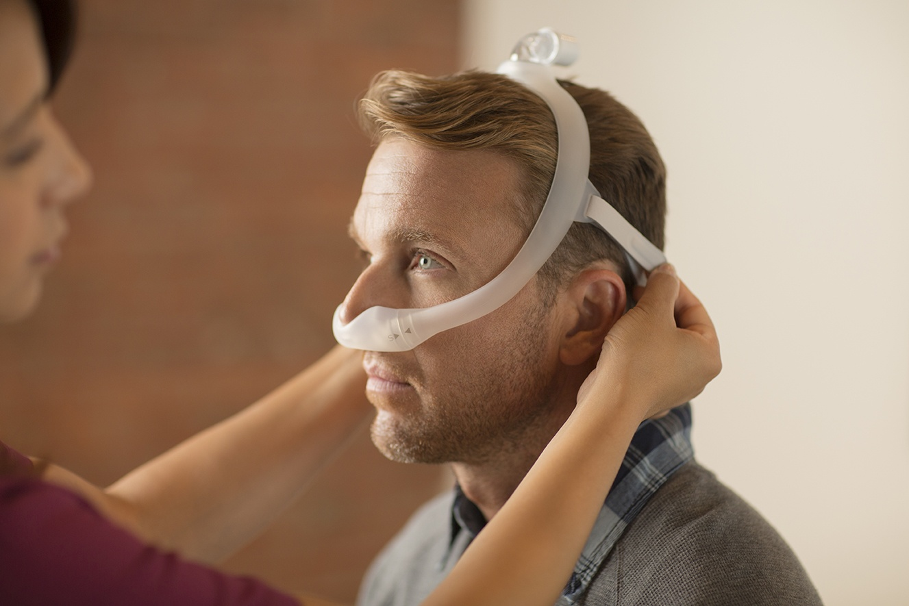 A woman is helping a man seated in a chair put on the Philips Respironics DreamWear nasal mask, which fits under the nose.