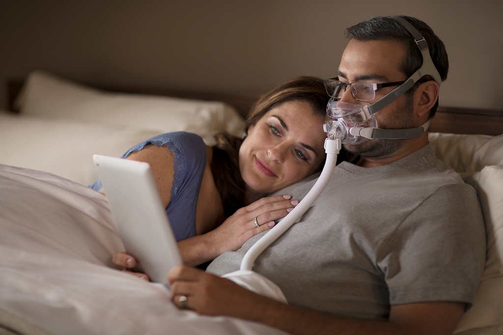 A man wearing a Philips Respironics Amara View Full Face Mask, reading glasses, and using his tablet with a woman in bed. 