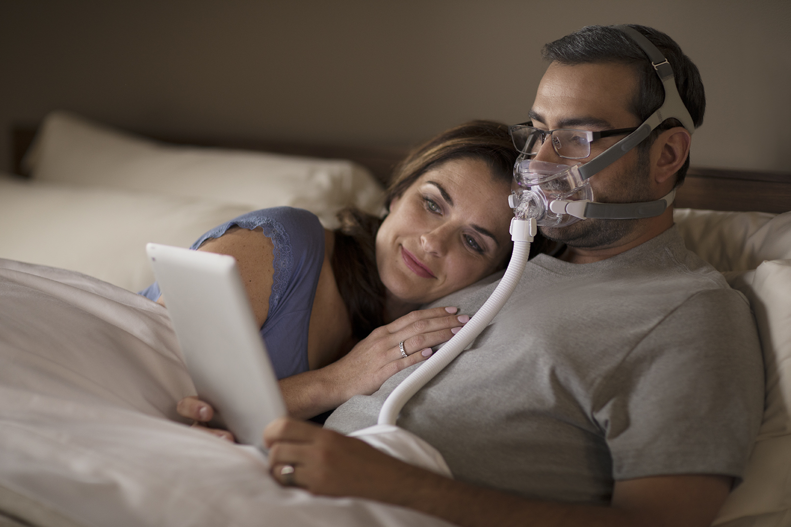A man wearing a Philips Respironics Amara View Full Face Mask, reading glasses, and using his tablet with a woman in bed. 