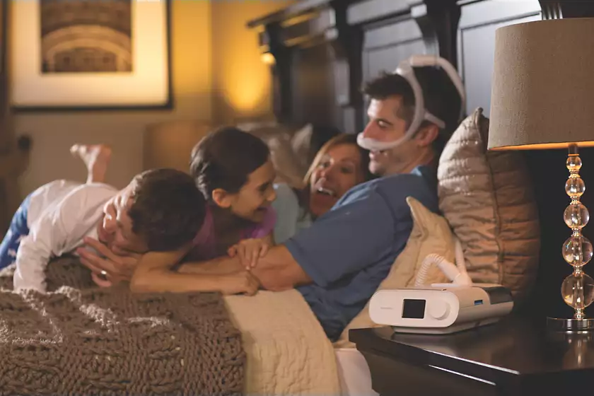 A Philips Respironics DreamStation Auto CPAP user with the machine by the bedside, wearing a DreamWear Nasal Mask, lying in bed with two kids and a woman.