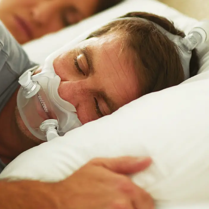 Close-up of a man wearing a Philips Respironics DreamWear Full Face CPAP mask connected to a CPAP machine, lying in bed.