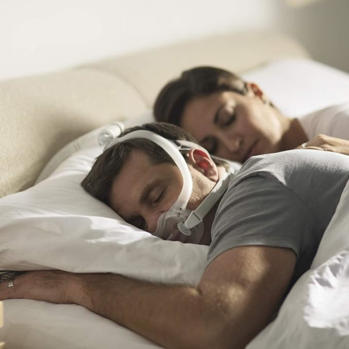Close-up of a man wearing a Philips Respironics DreamWear Full Face CPAP mask connected to a CPAP machine, lying in bed with a woman.
