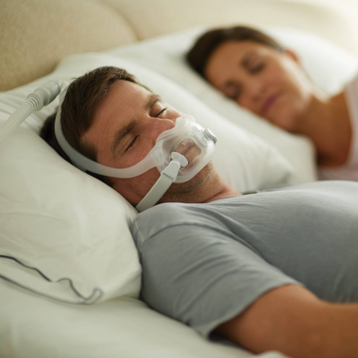 A man wearing a Philips Respironics DreamWear Full Face CPAP mask connected to a CPAP machine, lying in bed with a woman.