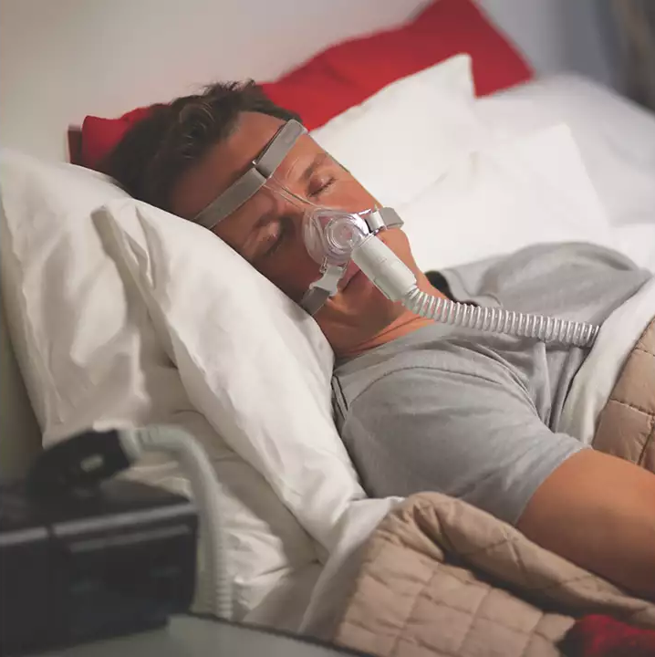 A man sleeping in bed, wearing the Philips Respironics Pico nasal mask connected to a CPAP device by the bedside.