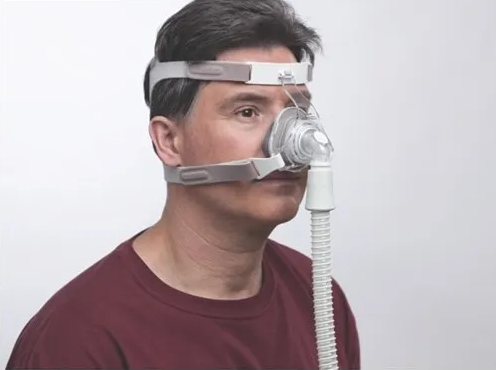A man wearing a Philips Respironics TrueBlue Nasal mask with headgear, connected to a CPAP machine.