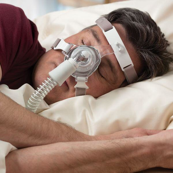 A man sleeping in bed, wearing a Philips Respironics TrueBlue Nasal mask connected to a CPAP machine.